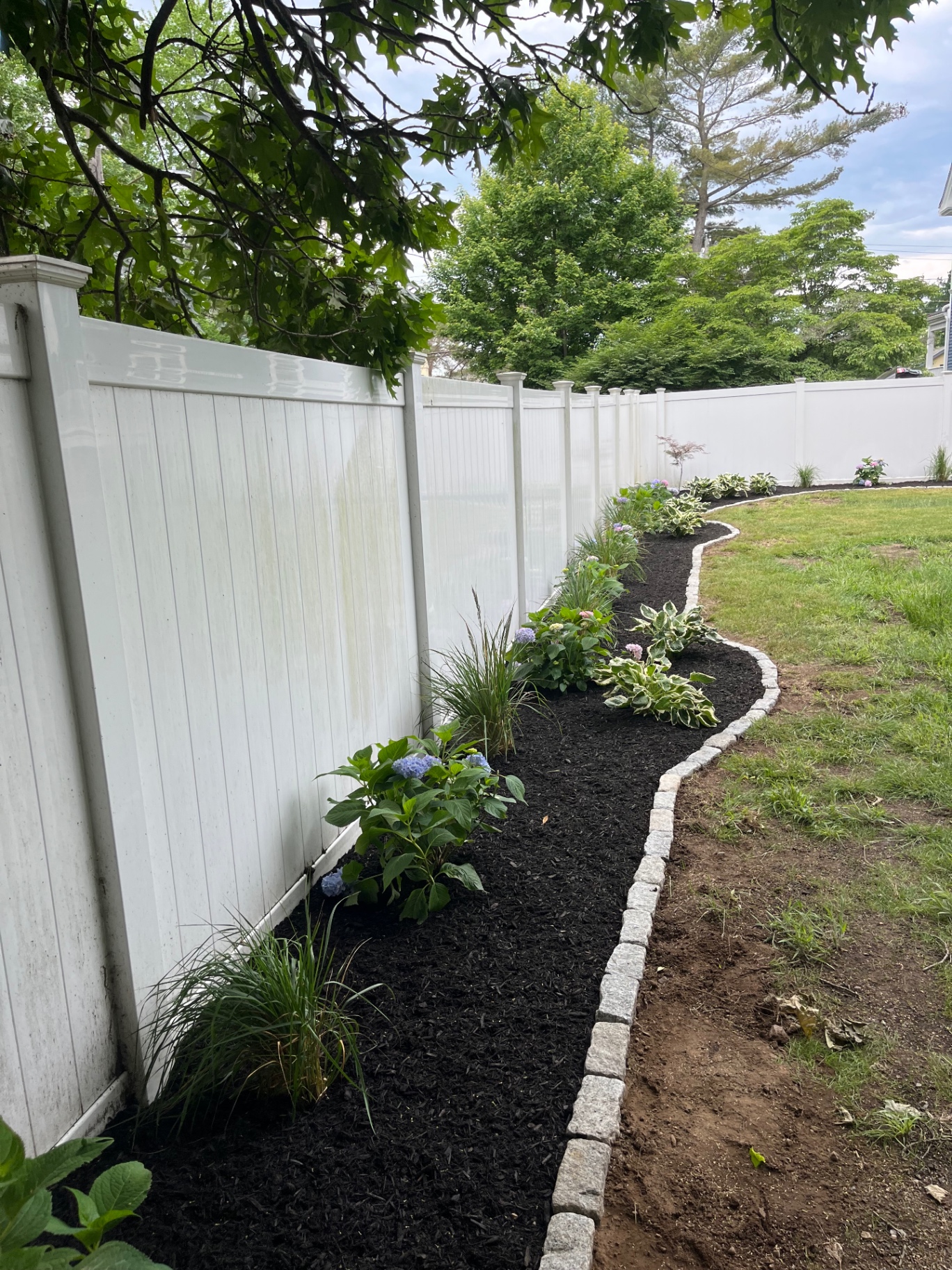 Long curved planting bed with cobblestone border along a white fence
